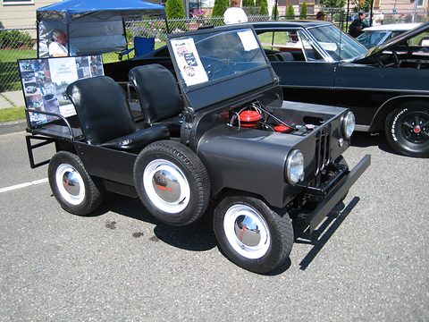 Small black open utility with twin rear wheels on each side, two seats and no bonnet, the engine visible, on a car park.