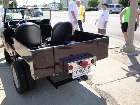 Rear of a small black open utility with two seats, a load box, a single tail lamp and a plate, parked on a pavement with people behind. Opens a larger view