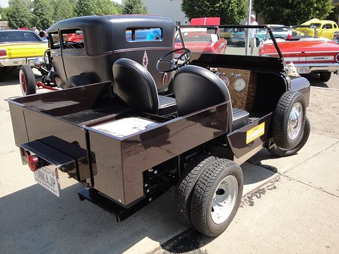 Rear quarter of a small black utility with a lengthened load bed, twin rear wheels and two seats, classic cars behind. Opens a larger view