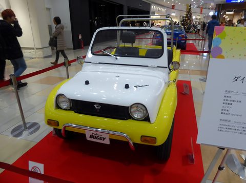 Yellow and white buggy on red carpet with a FELLOW BUGGY plate, shoppers walking past behind. Opens a larger view