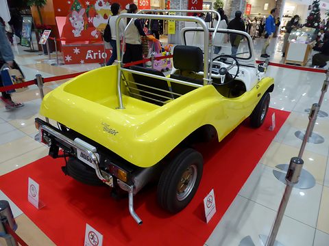 Rear three-quarter of a yellow open buggy with a roll bar, on red carpet behind a rope barrier. Opens a larger view