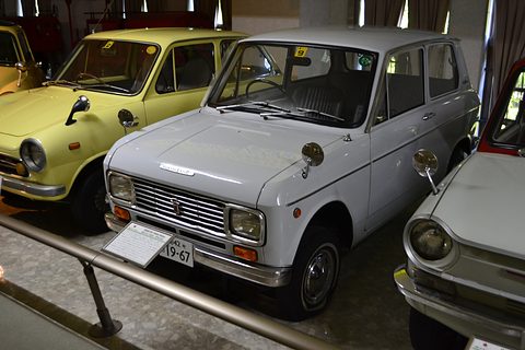 Pale grey boxy saloon behind a steel rail, a yellow small car to its left. Opens a larger view
