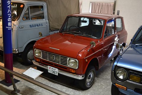 Red boxy saloon with a chrome grille and rectangular headlamps, a grey truck and blue sign behind. Opens a larger view