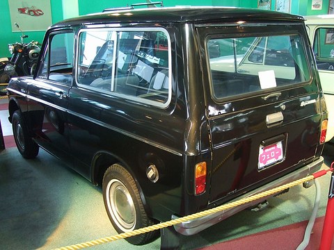 Rear three-quarter of a black boxy estate van behind a yellow rope, on a dark museum floor.