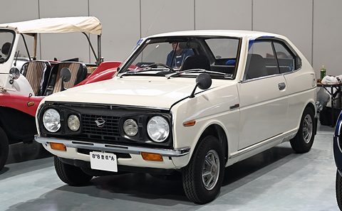 White two-door small saloon with four round headlamps in a black grille, a red buggy at left.