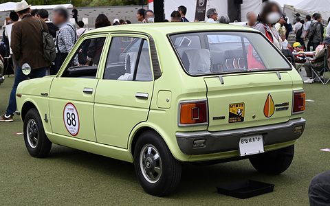 Rear three-quarter of a lime-green hatchback on grass, a crowd of visitors behind. Opens a larger view