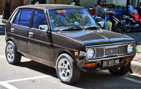 Dark brown four-door small hatchback parked on a street with motorcycles behind. Opens a larger view
