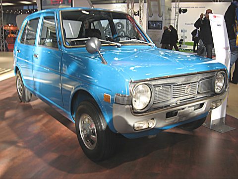 Light blue four-door small hatchback on a dark wooden turntable, a display stand behind. Opens a larger view