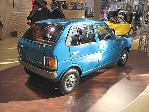 Rear three-quarter of a light blue four-door hatchback on a wooden stand, visitors behind. Opens a larger view