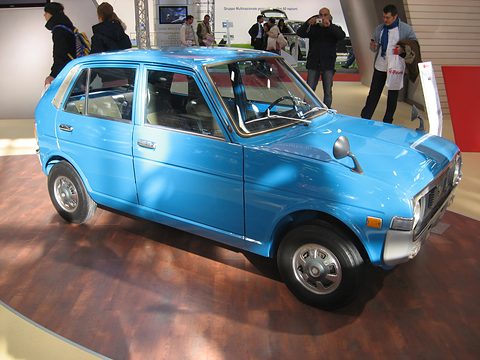 Light blue four-door hatchback from the front left on a wooden floor, visitors standing behind. Opens a larger view