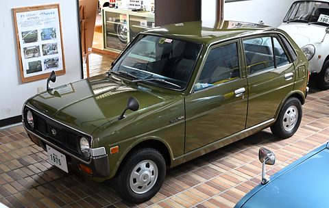 Olive green four-door hatchback with round headlamps on a tiled floor, a poster on the wall. Opens a larger view
