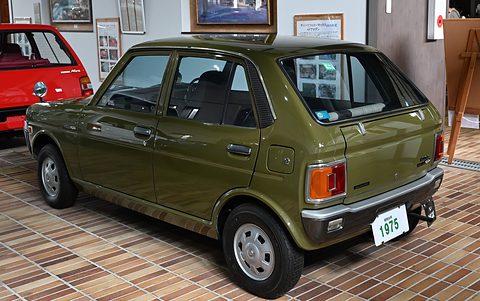 Rear three-quarter of an olive green four-door hatchback on a tiled floor, a red car at left. Opens a larger view