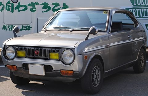 Grey pillarless coupé with a black grille, yellow fog lamps and round headlamps, a lettered van behind. Opens a larger view