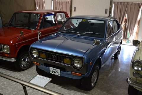 Metallic blue coupé on a museum floor beside a red saloon, behind a low rail. Opens a larger view