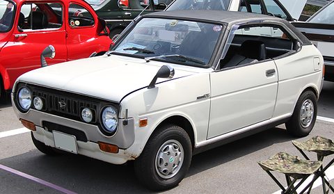 White coupé with a black roof, four round headlamps and wing mirrors, a camping stool beside it.