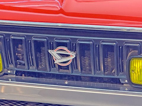 Close view of a black slatted grille with a chrome Daihatsu emblem and a yellow fog lamp, orange bonnet above. Opens a larger view