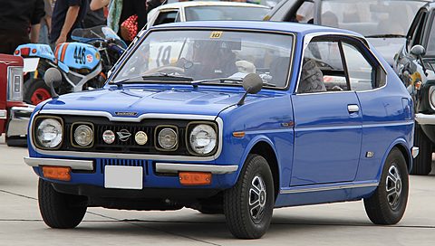 Bright blue coupé with four round headlamps in a black grille, other cars and visitors behind. Opens a larger view
