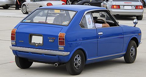 Rear three-quarter of a bright blue coupé with tall tail lamps, someone sitting in the driver’s seat. Opens a larger view