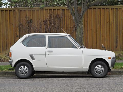Side view of a white two-door hatchback in front of a tall wooden fence and a tree. Opens a larger view