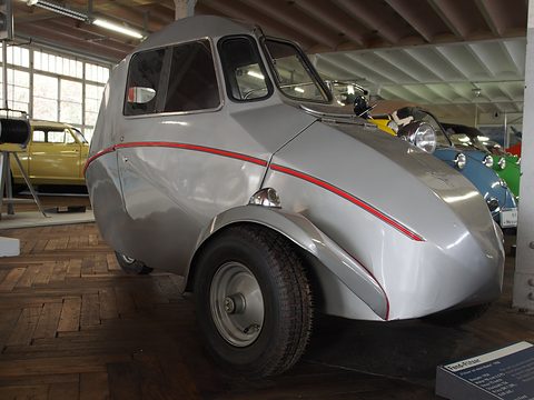 Silver teardrop-shaped single-seat three-wheeler with red side stripe and a large front wheel in its own wing, beside other microcars.
