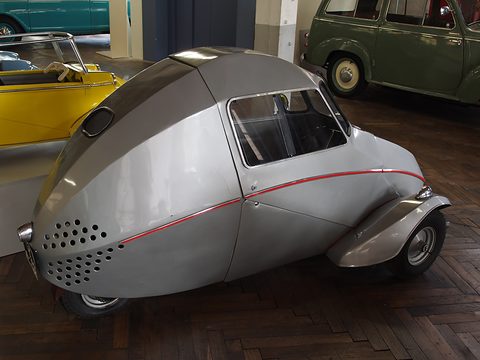 Rear three-quarter of a silver egg-shaped three-wheeler with rows of round vent holes, a small rear window and red pinstripe.