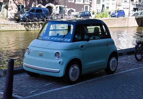 Mint-green rounded two-seater with a black roof parked on brick paving by a canal, houses and bicycles on the far bank. Opens a larger view