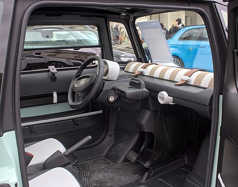 Dashboard of a small car with a two-spoke steering wheel and a striped roll along the dash, seen through the open door. Opens a larger view