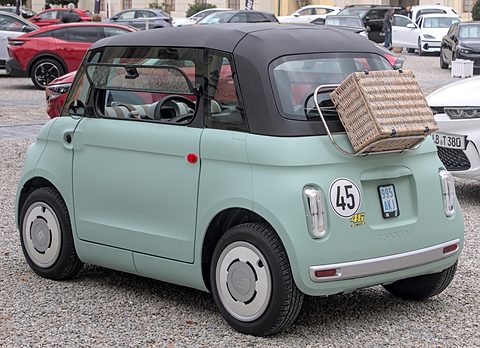 Rear of a pale green rounded two-seater with a picnic basket strapped to its rack and a 45 sticker, on gravel. Opens a larger view