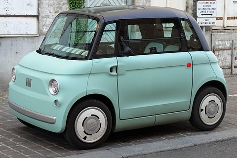 Pale green rounded two-seater with a black roof and white wheel covers parked on cobbles beside a white wall. Opens a larger view