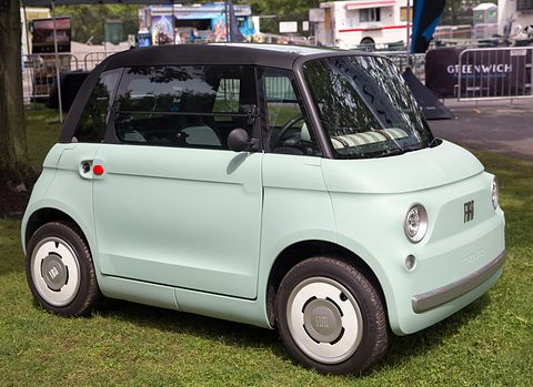 Pale green rounded two-seater with a black roof and white wheel covers, parked on grass with event stands behind. Opens a larger view