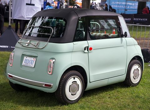 Rear of a pale green rounded two-seater with a chrome luggage rack and white wheel covers, on grass. Opens a larger view