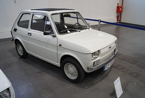 White small two-door car with a fabric sunroof and chrome bumpers on a grey show floor beside a placard. Opens a larger view