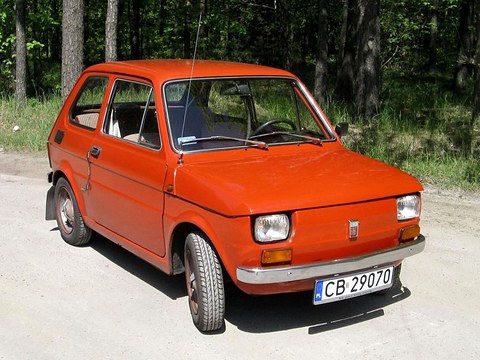 Orange-red small two-door car with chrome bumpers and a Polish plate on a sandy track in a pine wood. Opens a larger view