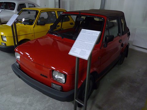 Red small car with a brown fabric hood and black bumpers beside a museum information stand, a yellow 126p behind. Opens a larger view
