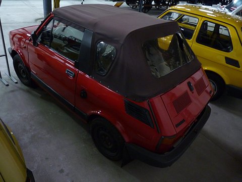 Rear three-quarter of a red small car with a brown fabric hood and slotted engine lid in a museum hall. Opens a larger view