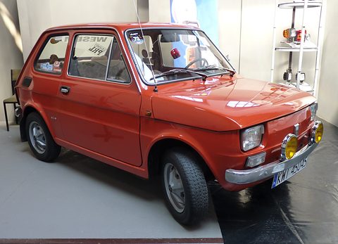 Red small two-door car with chrome bumpers and two yellow spotlamps on a grey show stand. Opens a larger view