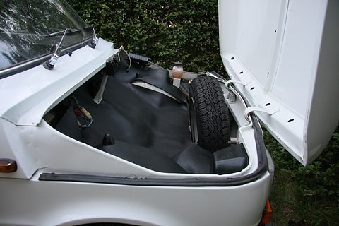 Open front lid of a white car showing a black-lined luggage well and an upright spare wheel. Opens a larger view