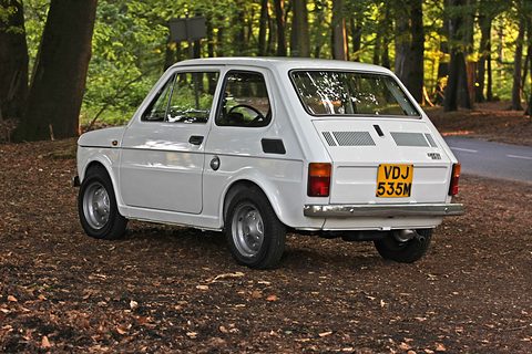 Rear three-quarter of a small white car with two sets of slots in its engine lid and a yellow UK plate, among trees. Opens a larger view