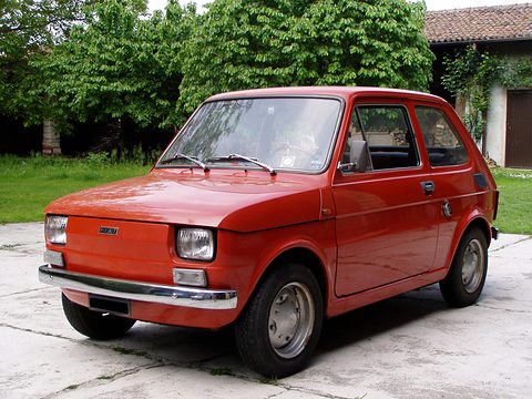 Small red two-door car with chrome bumpers on a concrete yard in front of trees and a tiled-roof building. Opens a larger view