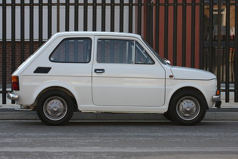 Side profile of a small white two-door car in front of black railings and a brick building. Opens a larger view