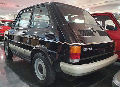 Rear three-quarter of a black small car with a gold side stripe, beige bumpers and square tail lamps on a shiny floor. Opens a larger view