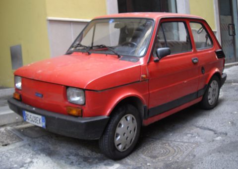 Faded red small hatchback with black plastic bumpers and a side strip parked in front of a yellow wall. Opens a larger view