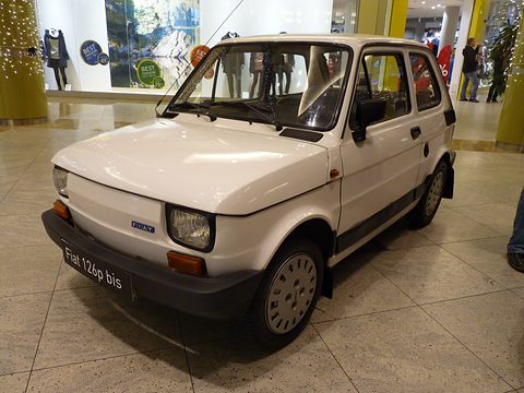 White small hatchback with black bumpers and a ‘Fiat 126p bis’ plate on a tiled mall floor, shop windows behind. Opens a larger view