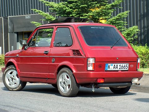 Rear three-quarter of a red small hatchback with a large rear window, white alloy wheels and a German plate. Opens a larger view