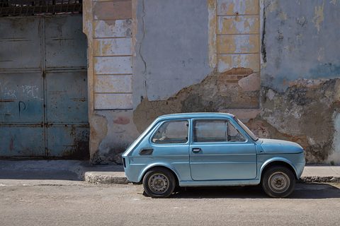 Side view of a pale blue small car parked at a kerb in front of a crumbling painted wall and a steel door. Opens a larger view