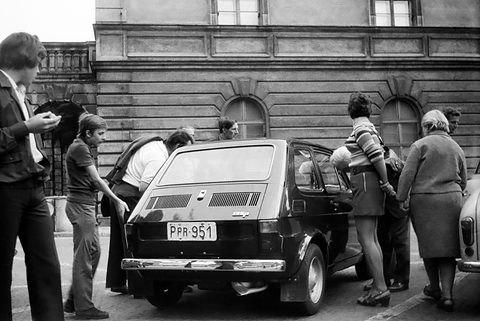 Rear view of a small dark two-door car with louvred engine lid, people leaning in at its open door and crowding round.