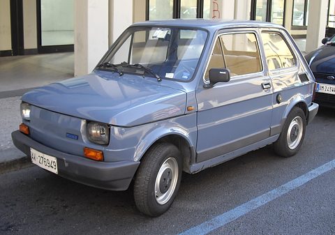 Blue-grey small two-door car with black plastic bumpers and a FIAT badge parked at a kerb beside a parking line. Opens a larger view