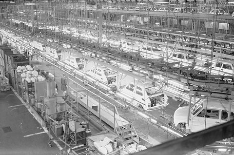 Black-and-white high view of a long line of car bodies hanging on conveyors in a factory, crates on the floor. Opens a larger view