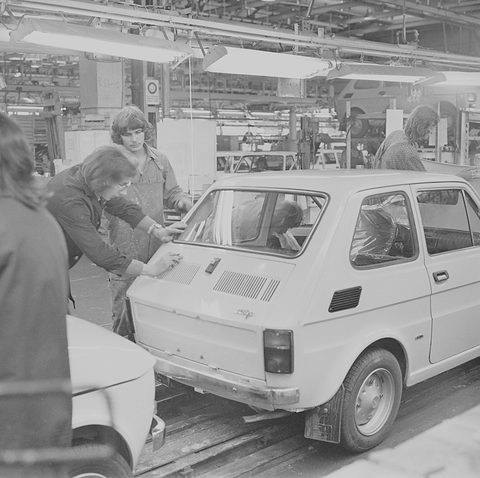 Black-and-white photograph of two young women leaning over the rear of a small car on a line, others working behind. Opens a larger view