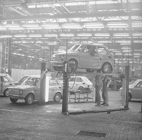 Black-and-white factory hall with a small car raised on a lift, workers beneath it and finished cars around.